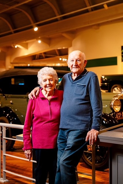 John Henry and Beatrice Marie “Peggy” Rendzio Man in blue shirt and blue jeans leans on barrier with his arm around woman in pink shirt and black pants in front of car display