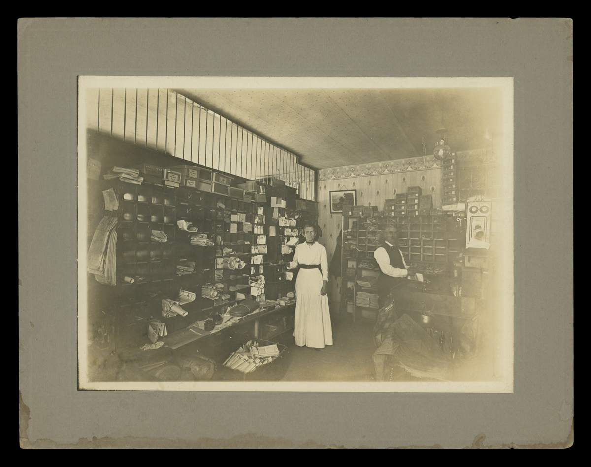 Postmaster Madison Gallogly and Mary Gallogly in the Hoxie, Kansas Post Office, circa 1913 Matted black-and-white photo of a man and woman in a room filled with mail bins and cubbies with items sorted into them