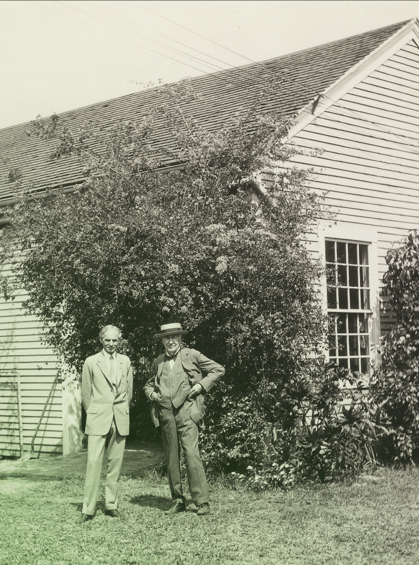Henry Ford and Thomas Edison with Fort Myers Laboratory at Its Original Site, Fort Myers, Florida, circa 1925 Two men in suits, one wearing a hat, stand in front of a wooden building partially obscured by vegetation