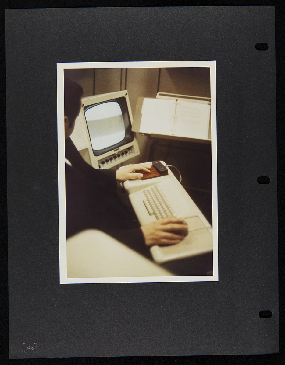 Page from Report on the SRI Fall Joint Computer Conference, December 27, 1968 Photo pasted onto black three-ring binder sheet depicting a person in front of a keyboard with a monitor to the side and copystand in front of them