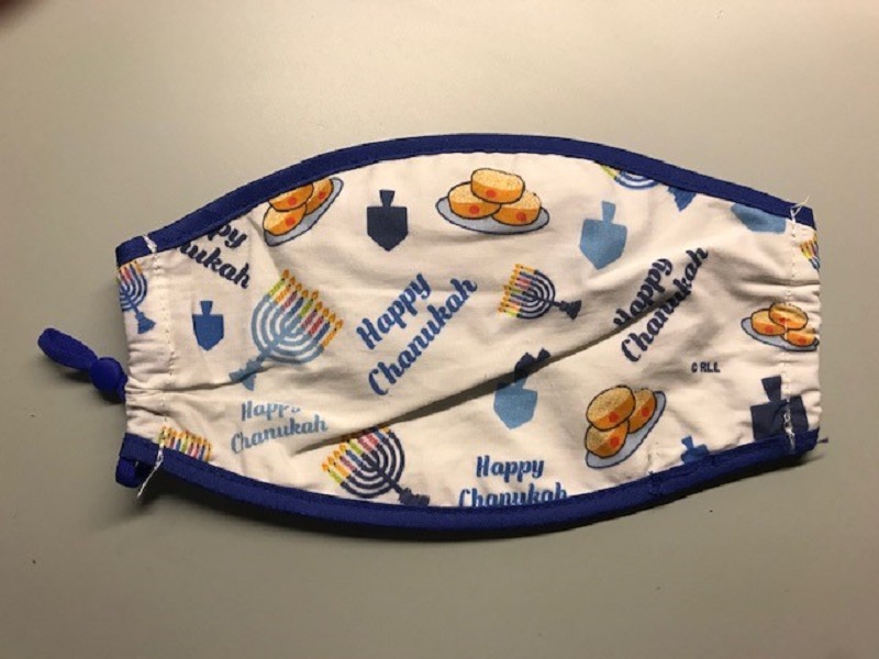 Beige facemask with dark blue trim and images of dreidels, menorahs, and jelly doughnuts, along with text