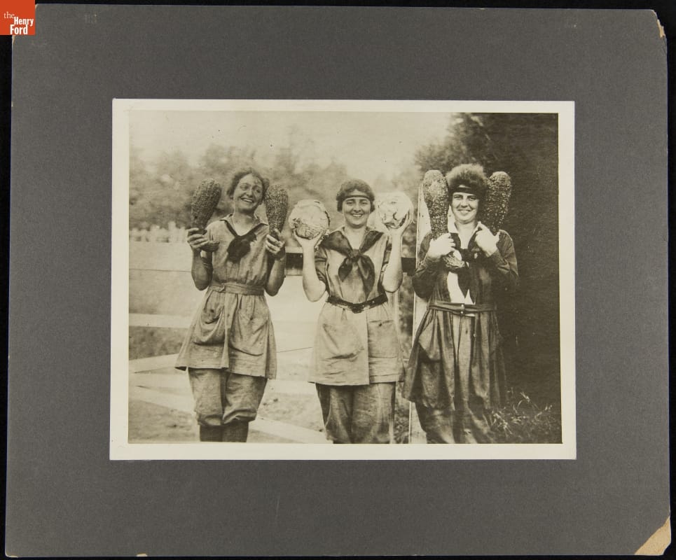 Black-and-white image of women, perhaps in some type of uniform, holding vegetables up in each hand