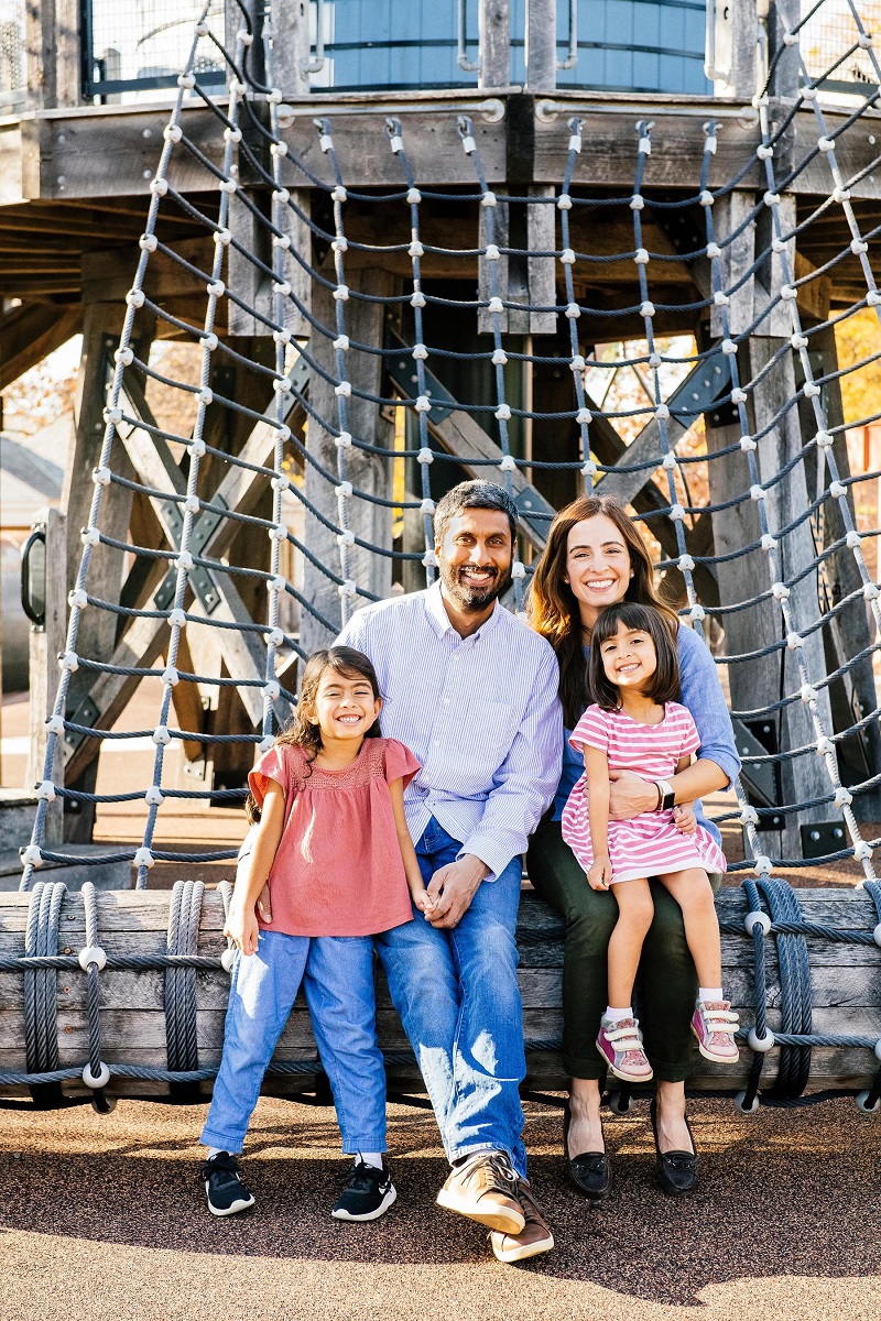 Man, woman, and two young girls sit and stand on a log in front of netting attached to a playground structure