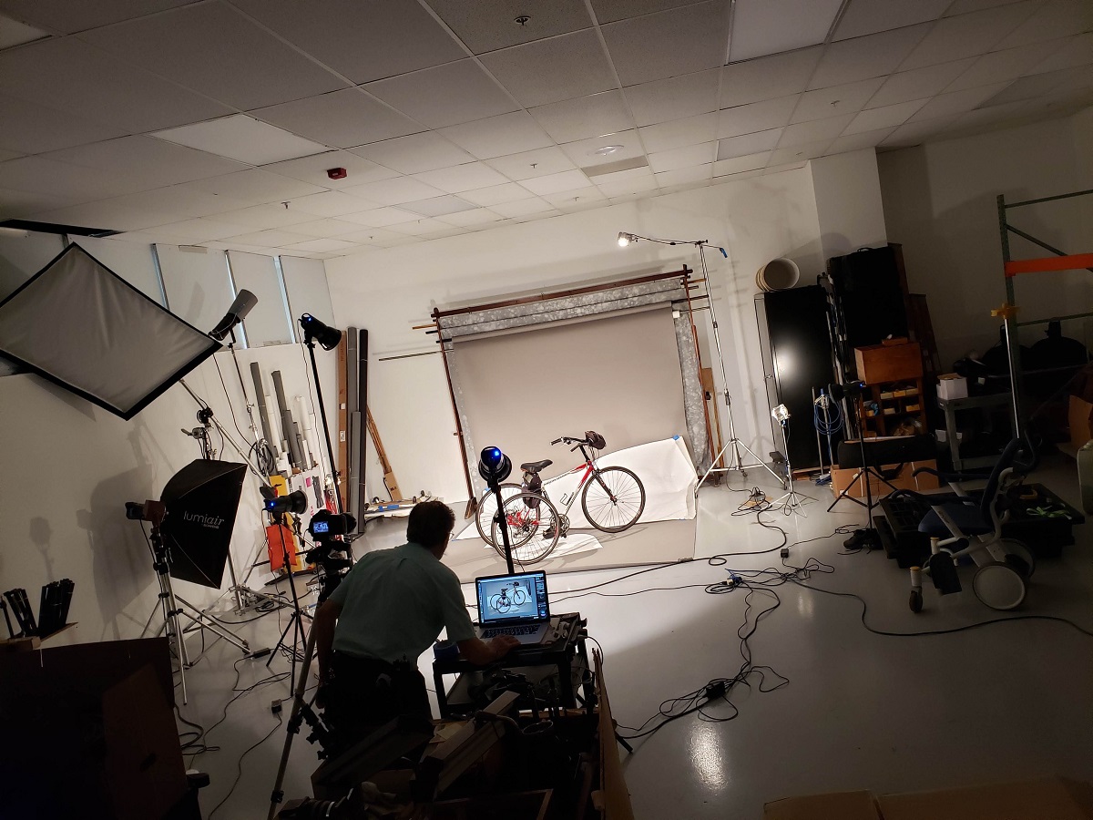 Setting up for the tricycle photograph Man stands at a laptop on a cart in a photo studio with a tricycle on the set