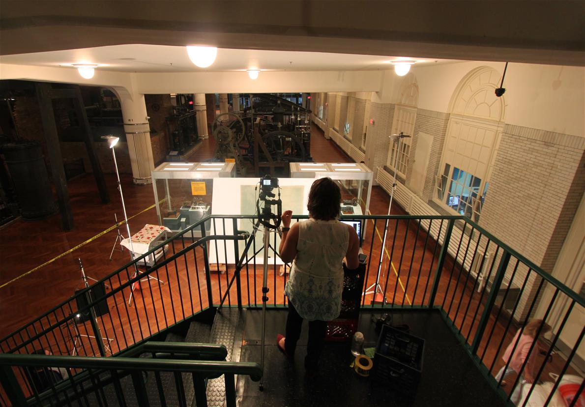 Getting ready to photograph quilts from the Highland Park Engine catwalk in Henry Ford Museum of American Innovation. Person with camera on landing at top of staircase, looking down over railing toward a large tilted board with a textile on it