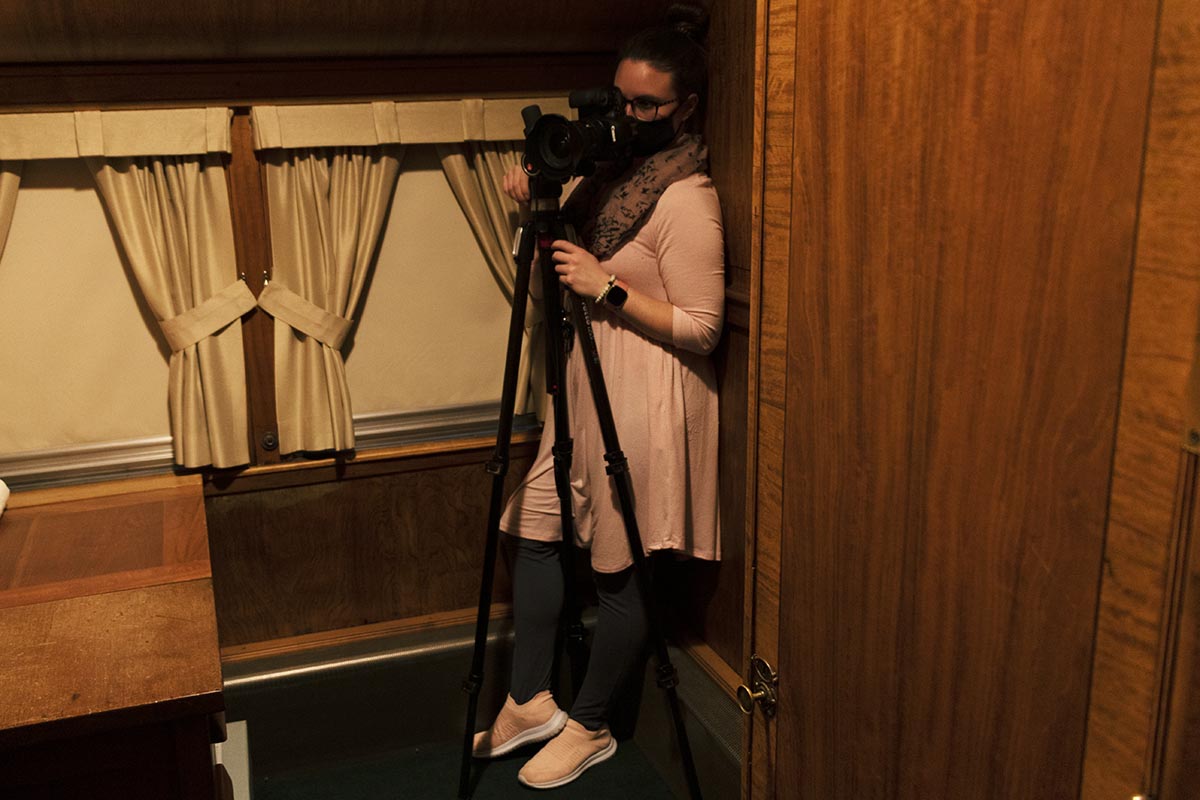 Digital Imaging Specialist Jillian Ferraiuolo, setting up the shot of the Fair Lane railroad car office Woman stands in tight corner of wood-paneled room behind a camera on a tripod