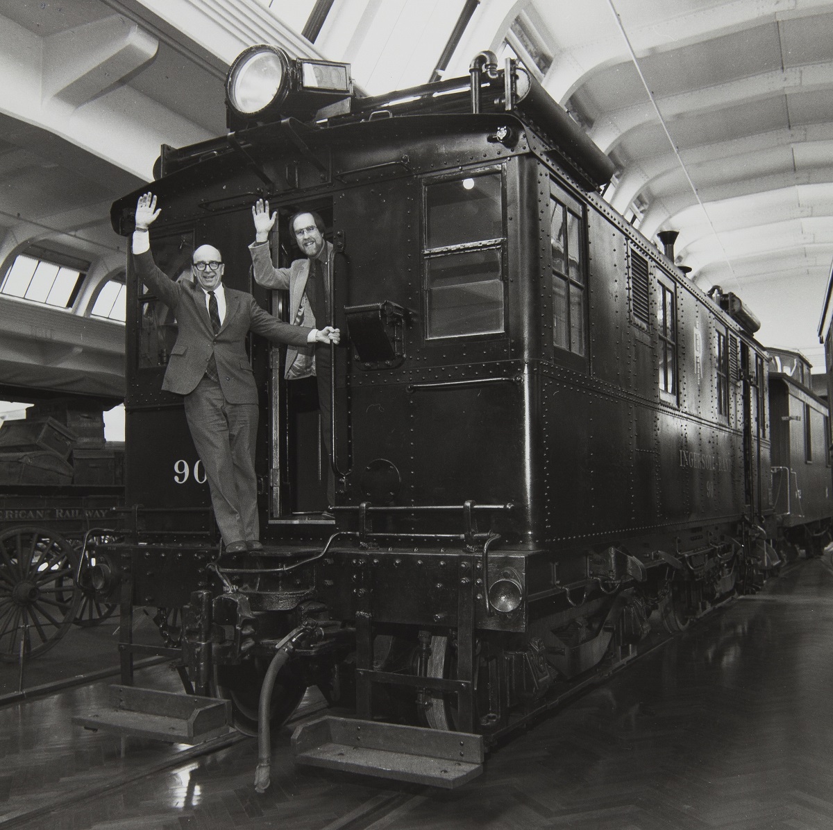 M. William Grant and Randy Mason with Restored Ingersoll-Rand Diesel-Electric Locomotive No. 90 in Henry Ford Museum, January 1985 Two men wave from the back of a rail car on tracks in a large room