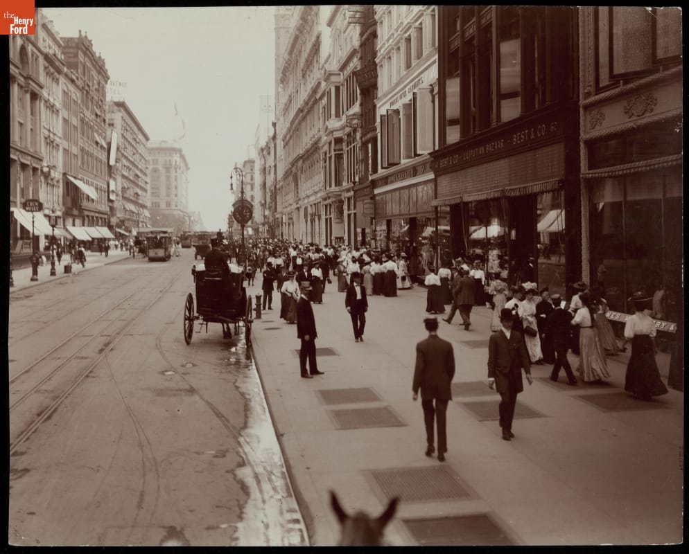 "West 23rd Street, New York City," circa 1908 Black-and-white photo of city street with tall buildings and wide sidewalks crowded with people; carriages and streetcars visible on street