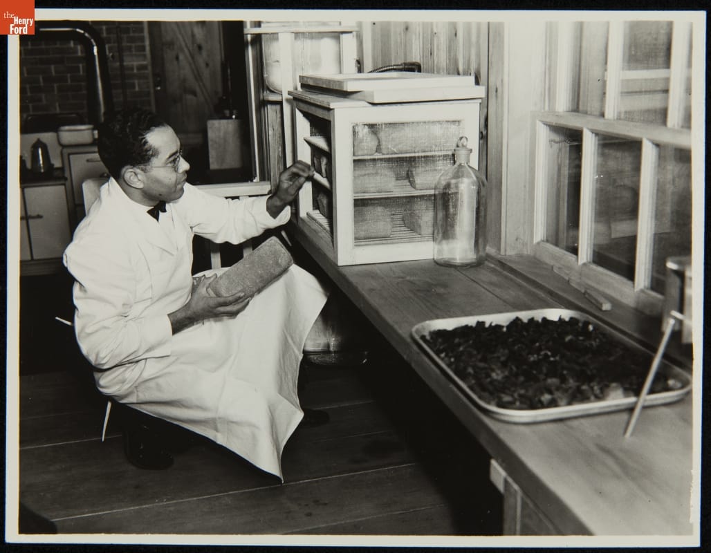Worker Storing Bread in the Soybean Laboratory in Greenfield Village, Dearborn, Michigan, circa 1935 Man in white apron kneels with loaf of bread in his hand before small cabinet filled with loaves of bread