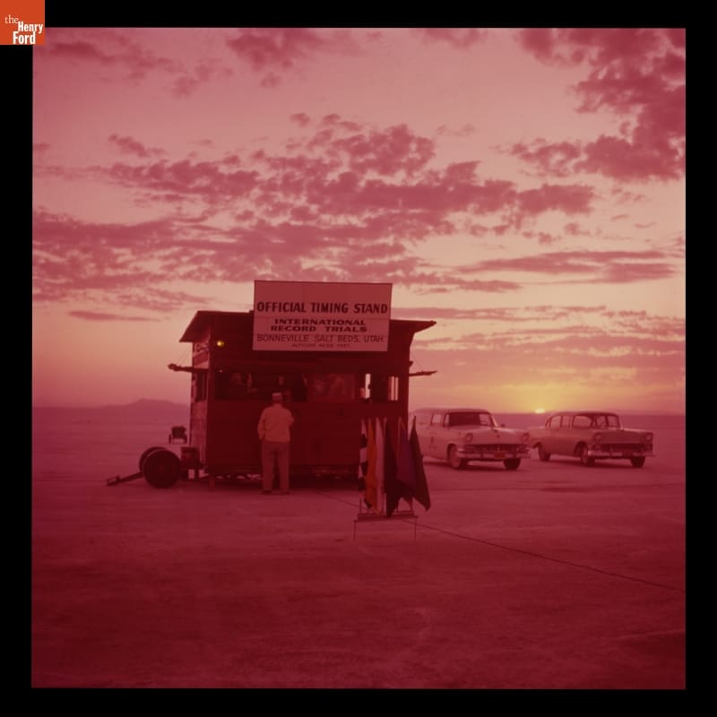 Pink-tinted photo showing shack, people, cars in a large open expanse