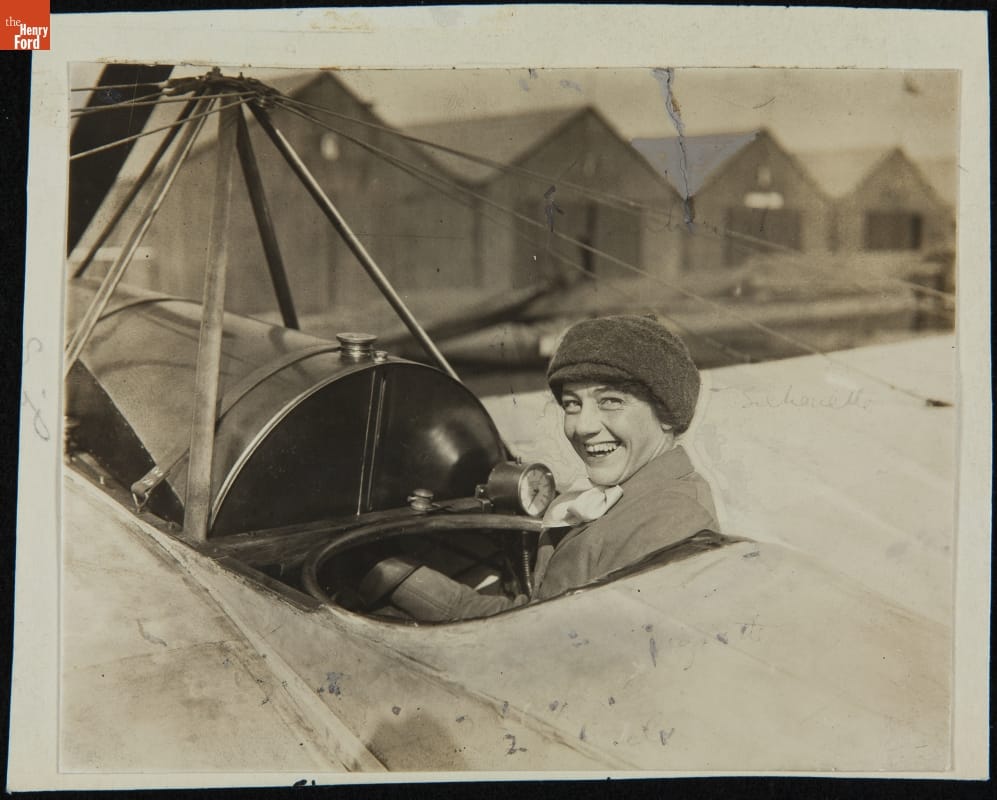Agnes Firth in a Caudron Airplane, 1911-1912 Woman in an open plane cockpit smiles at the camera; a row of sheds is in the background