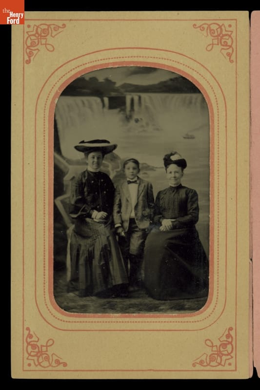 Clara Ford, Edsel Ford and Martha Bryant in a Photographer's Studio Portrait Taken at Niagara Falls, 1901 Two seated women in dresses and hats with small boy standing between them, all in front of a painted backdrop of a waterfall