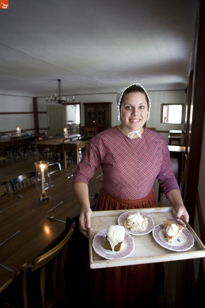 Smiling woman in pink dress and white bonnet holds a tray of food in a large room with low ceilings and long wooden tables