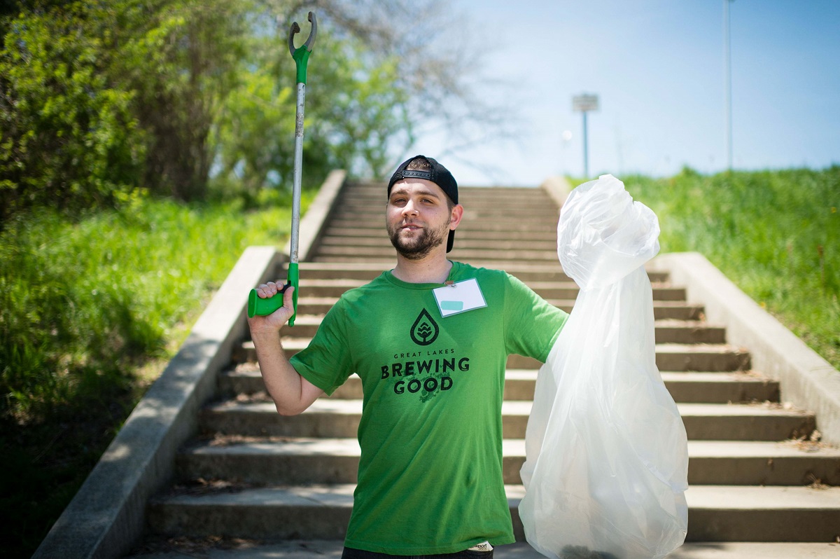 A Brewing Good community clean-up effort by Great Lakes Brewing Co. Man in baseball cap and green t-shirt holds up grabber and trash bag while standing on concrete steps with trees and grass on either side