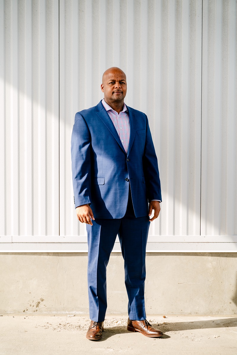 Medium-skin tone man in a blue suit stands in front of a corrugated white metal wall 