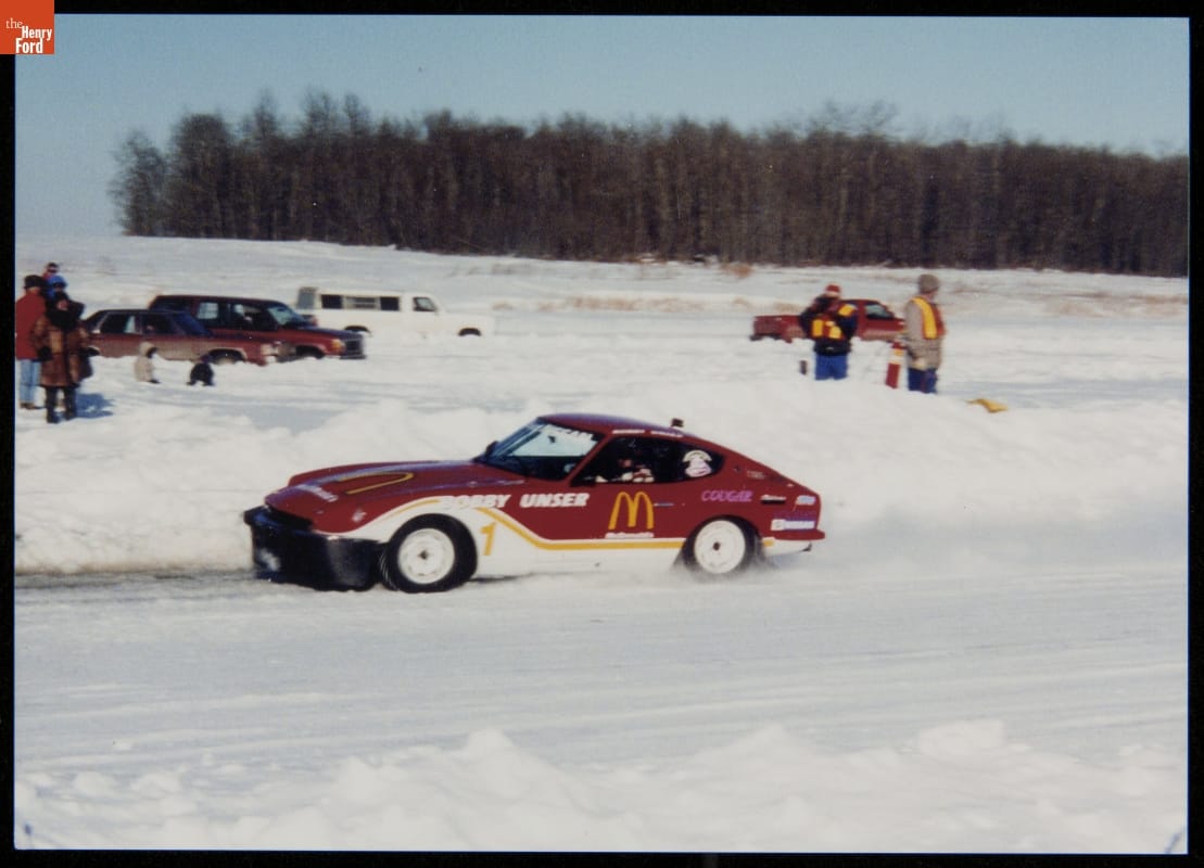 Red and white race car on a snowy road with snowbanks on either side, with people, other cars, and trees in background