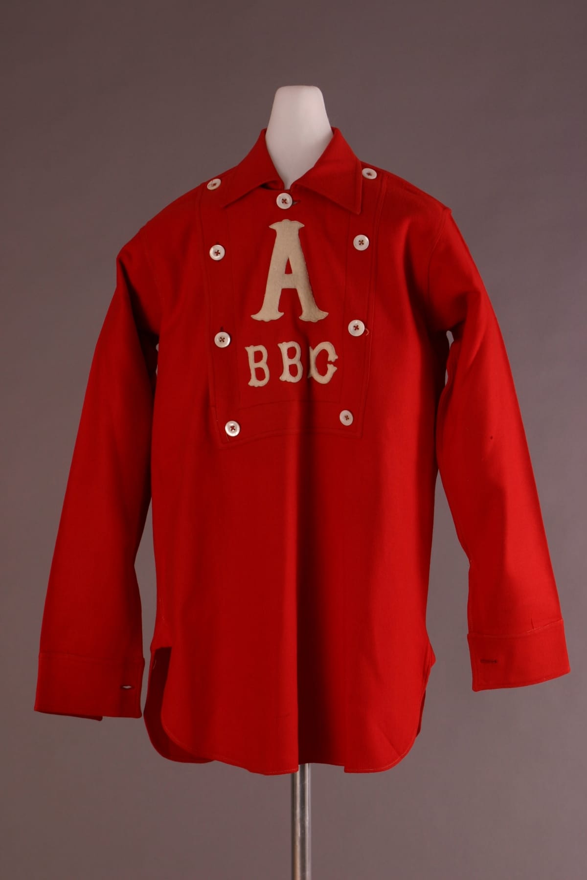 Baseball Uniform Shirt, 1865-1885 Red shirt with collar and white buttons around the placket, featuring white letter "A" followed by "BBC"