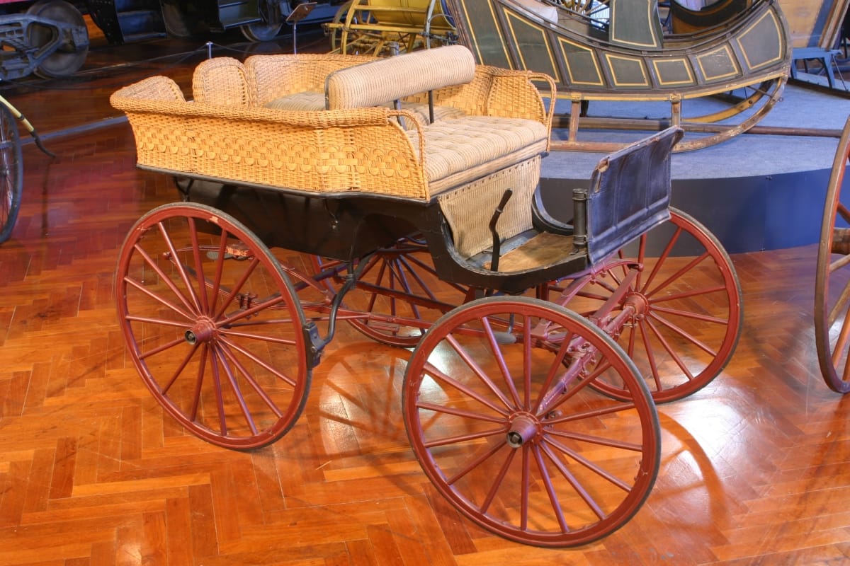 Pony Wagonette, circa 1900 Black open carriage with red wheels and beige rattan (?) sides, displayed in a room with other vehicles