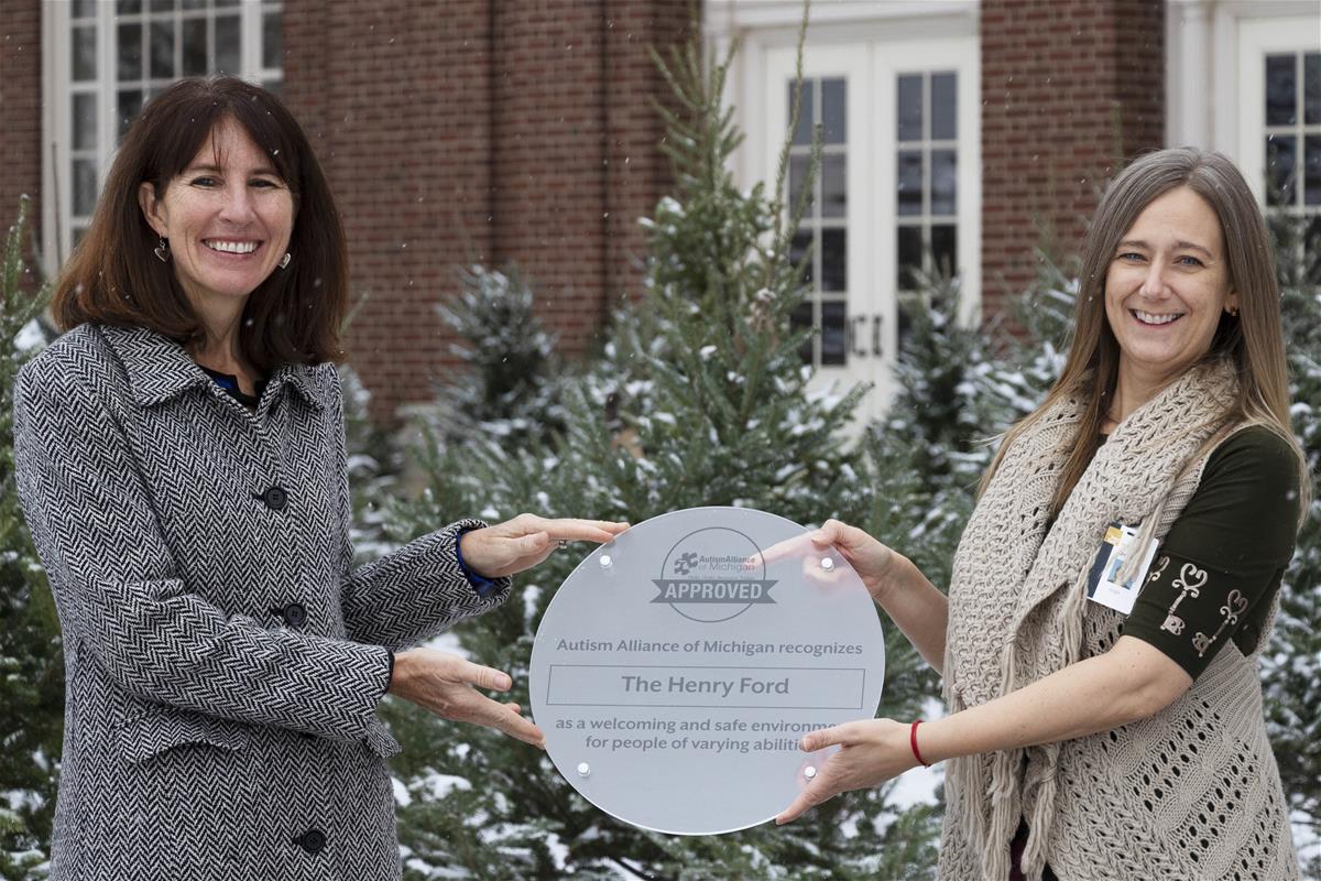 The Henry Ford receives AAoM's Seal of Approval Two women hold a frosted plastic plaque with text in front of snow-covered evergreens and a brick building