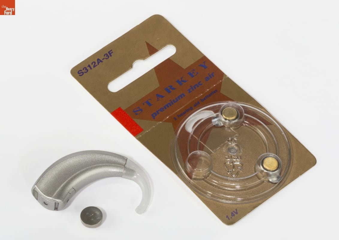 Starkey Laboratories S Series Behind-the-Ear Hearing Aid, 2010 Silver and white comma-shaped device, coin battery, and cardboard and plastic blister packaging containing text