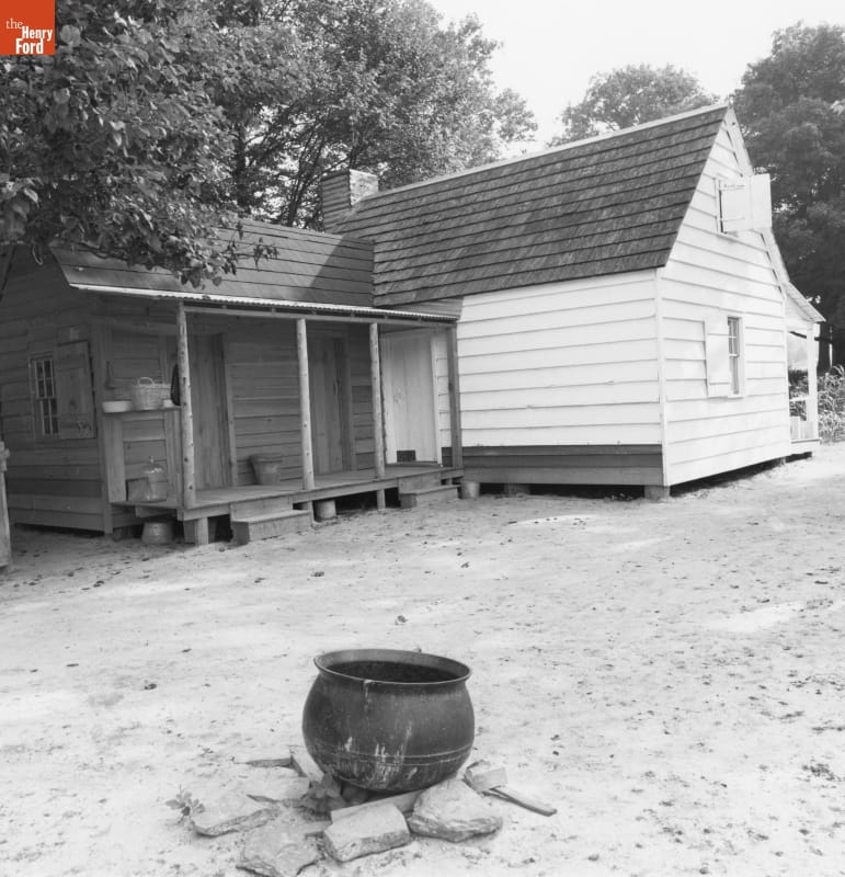 Mattox Family Home in Greenfield Village, 1991 Black-and-white photo of wooden house with large iron kettle over ring of stones in dirt yard