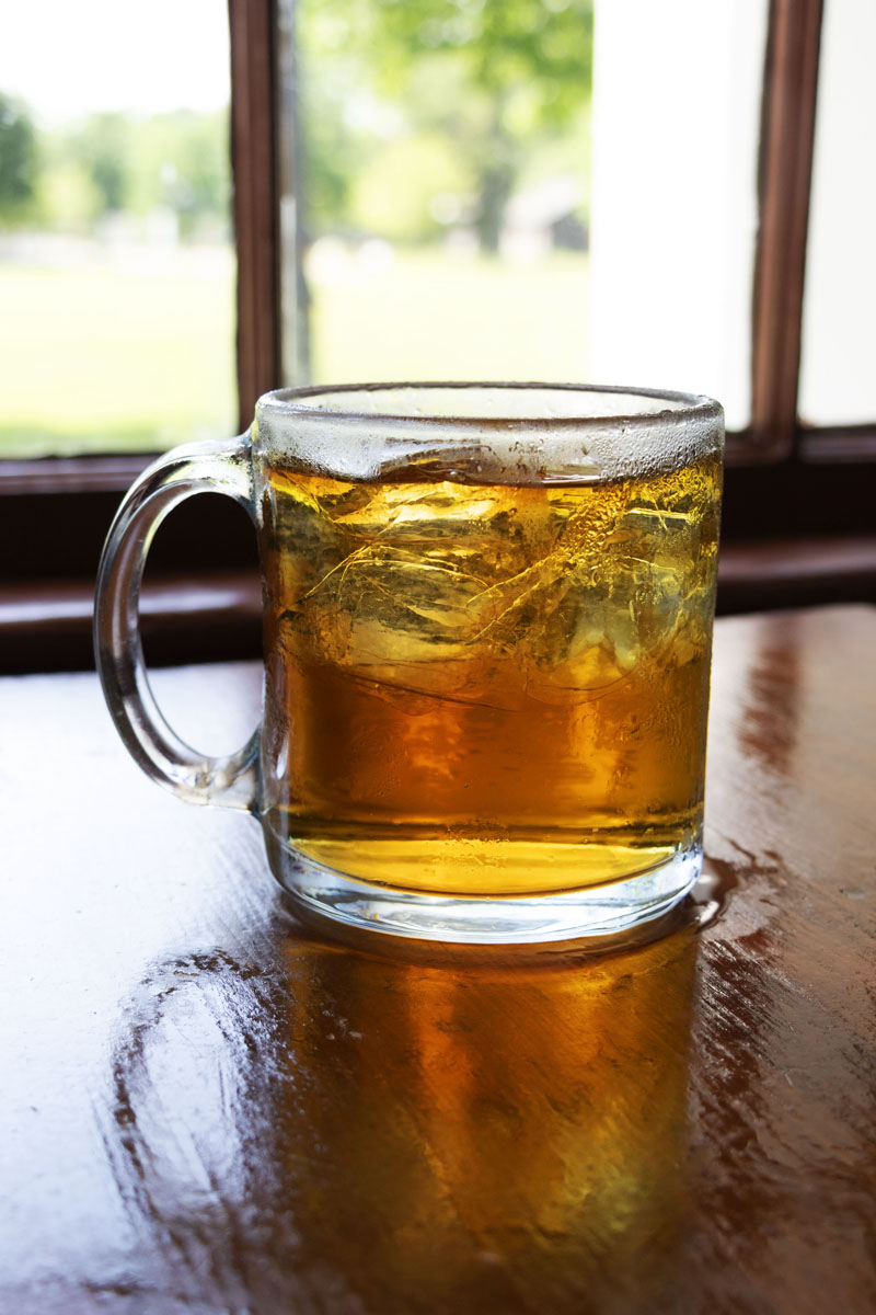 Eagle Tavern's Stone Wall Clear glass mug with amber liquid and ice inside, sitting on wooden table in front of window