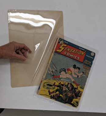 Comic book being bagged, boarded, and encapsulated for storage, display, or handling Hand sliding comic book onto a board with plastic over top