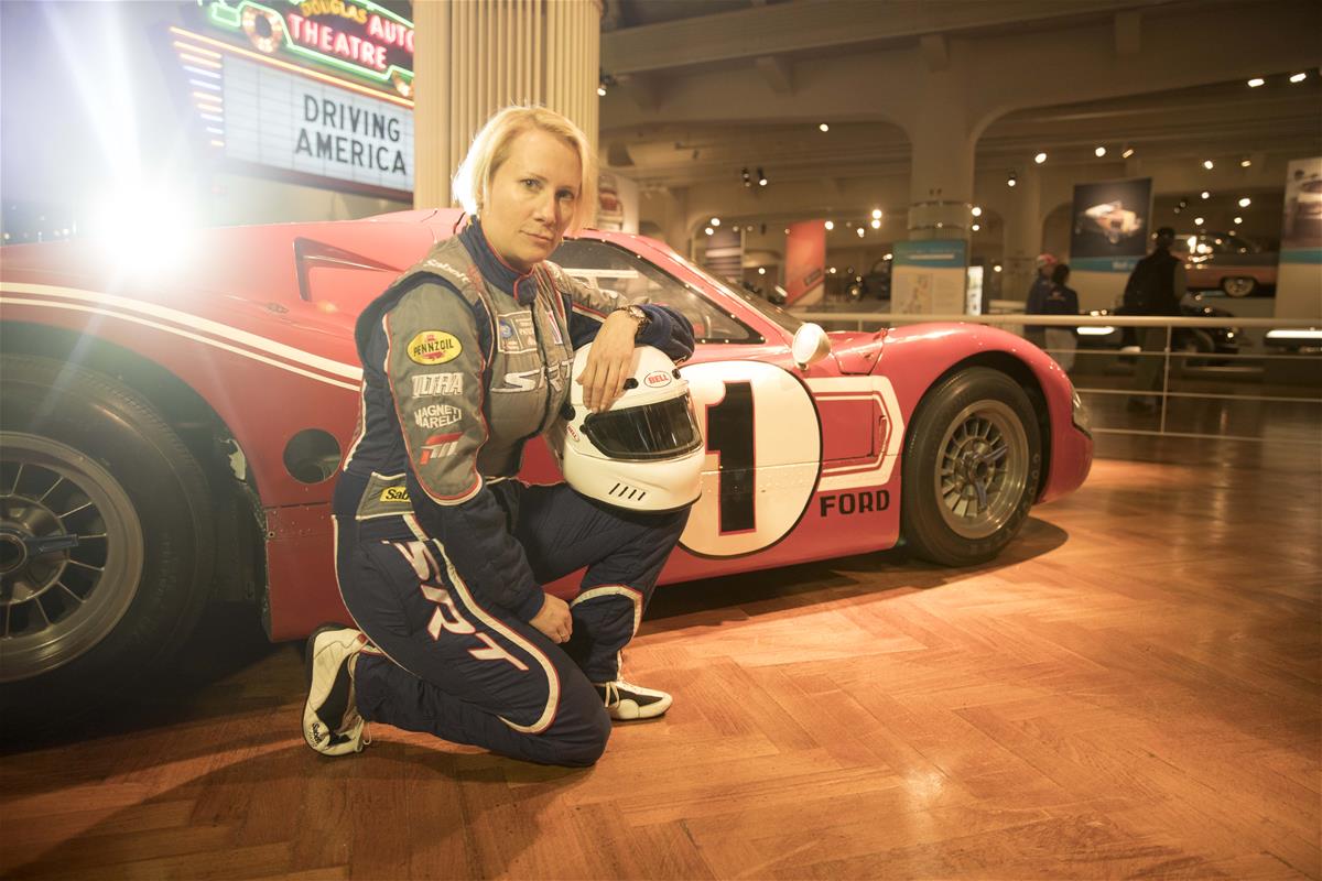 Beth Paretta poses with the 1967 Ford Mark IV Race Car at The Henry Ford in 2018 Woman in jumpsuit holding a helmet kneels on wood floor in a building next to low red race car with dramatic backlighting