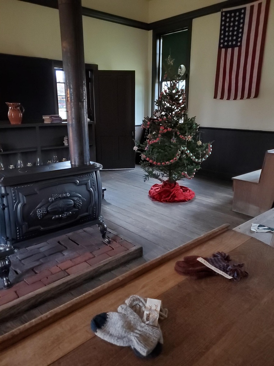Scotch Settlement School during Holiday Nights in Greenfield Village Room interior containing Christmas tree, wood-burning stove, bookshelves, and American flag hanging from wall