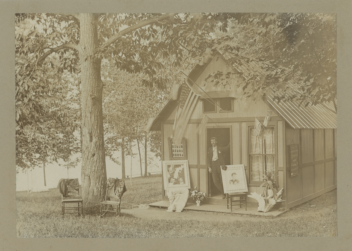 Photographer outside His Tintype Studio, circa 1890 Man leaning in doorway of small wooden building in wooded location; chairs and portraits outside
