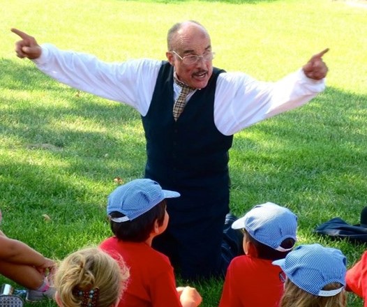 Anthony Lucas at Discovery Camp at The Henry Ford Man in suit (vest, pants, and white shirt) kneels on the ground with hands outstretched while children in baseball caps watch