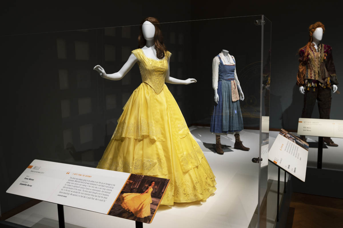 Costumes from the live-action movie Beauty and the Beast in the Heroes and Villains: The Art of the Disney Costume exhibit Display case containing mannequins wearing a yellow dress, simple blue jumper, and pants with elaborate jacket; large labels are posted in front of the display
