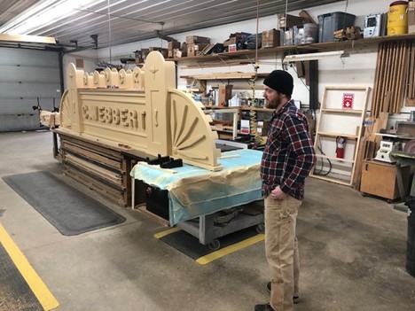 Carpenter Mike Zemney with the nearly completed C. Webbert Block sign he built. Man in knit hat, khakis, and plaid shirt stands next to large sign in workshop space