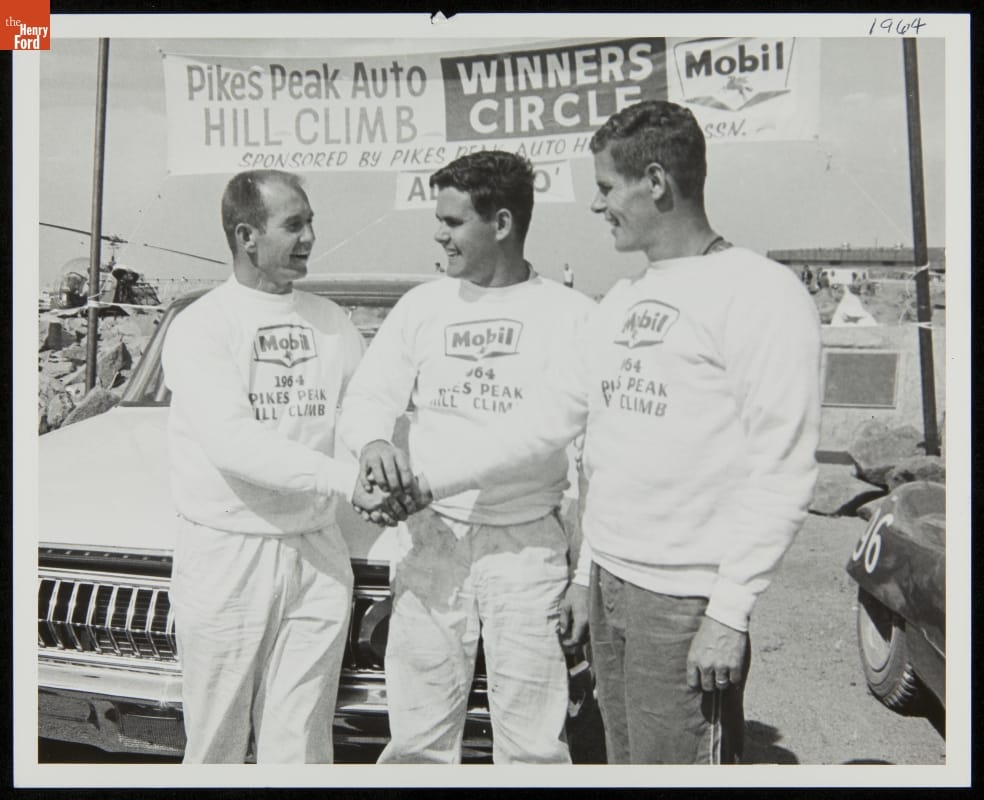 Wes Vandervoort, Al Unser, Sr., and Bobby Unser at Pikes Peak, 1964 Black-and-white photo of three men in matching sweatshirts joining hands in front of a car and banner