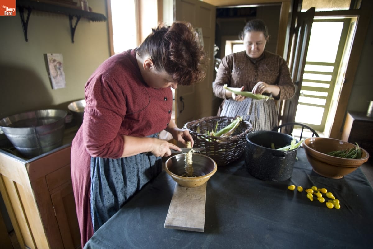 Firestone Farm in Greenfield Village, September 2007 / Photographed by Michelle Andonian Two women shuck and remove kernels from corn cobs at a kitchen table containing other dishes and food