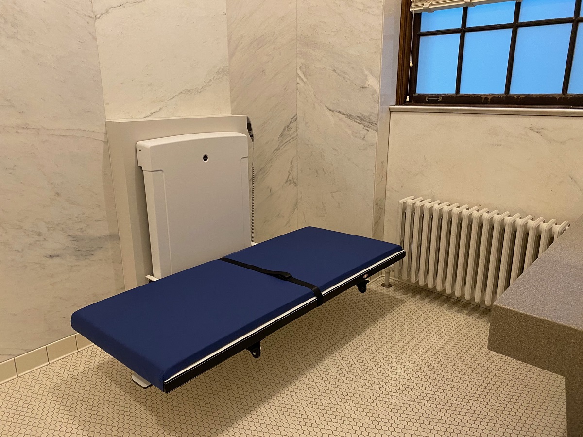 Adult changing table in one of our two new accessible companion care restrooms Blue padded table with machinery behind it in a space with tile floor and marble walls (also a window and radiator)