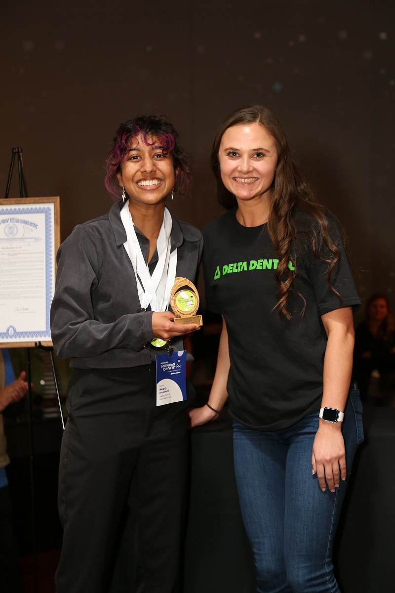 Meera R., the grand prize winner of Invention Convention Michigan 2022 A girl in a black shirt and top holds an award and smiles for the camera next to a woman in a black t-shirt and jeans