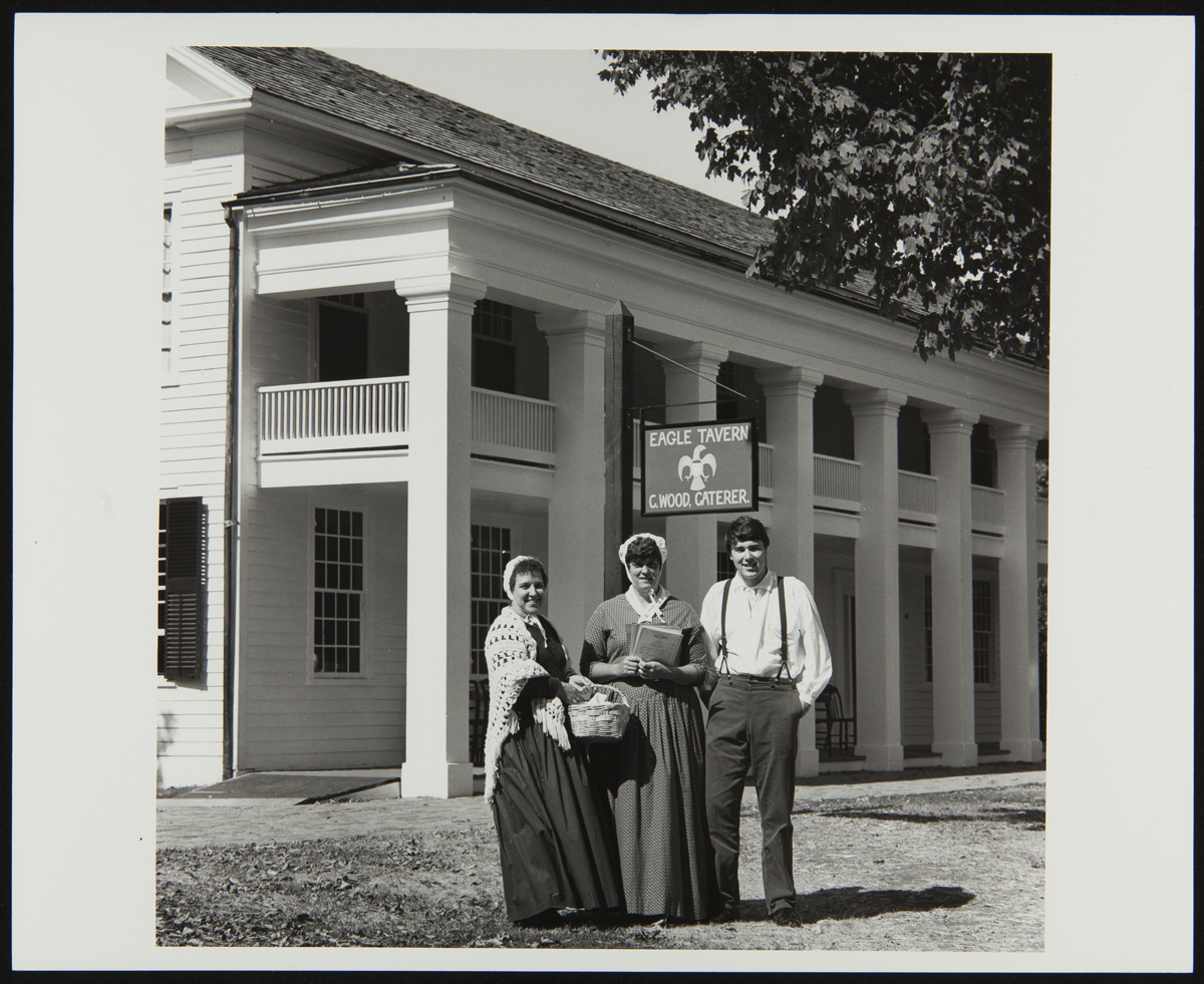 Historical Presenters outside Eagle Tavern in Greenfield Village, 1982 Three people in historical garments stand outside a two-story wooden building with sign containing text
