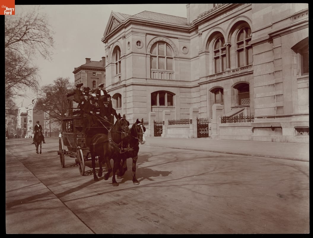 Two horses pull a large carriage loaded with people along a street lined with large buildings