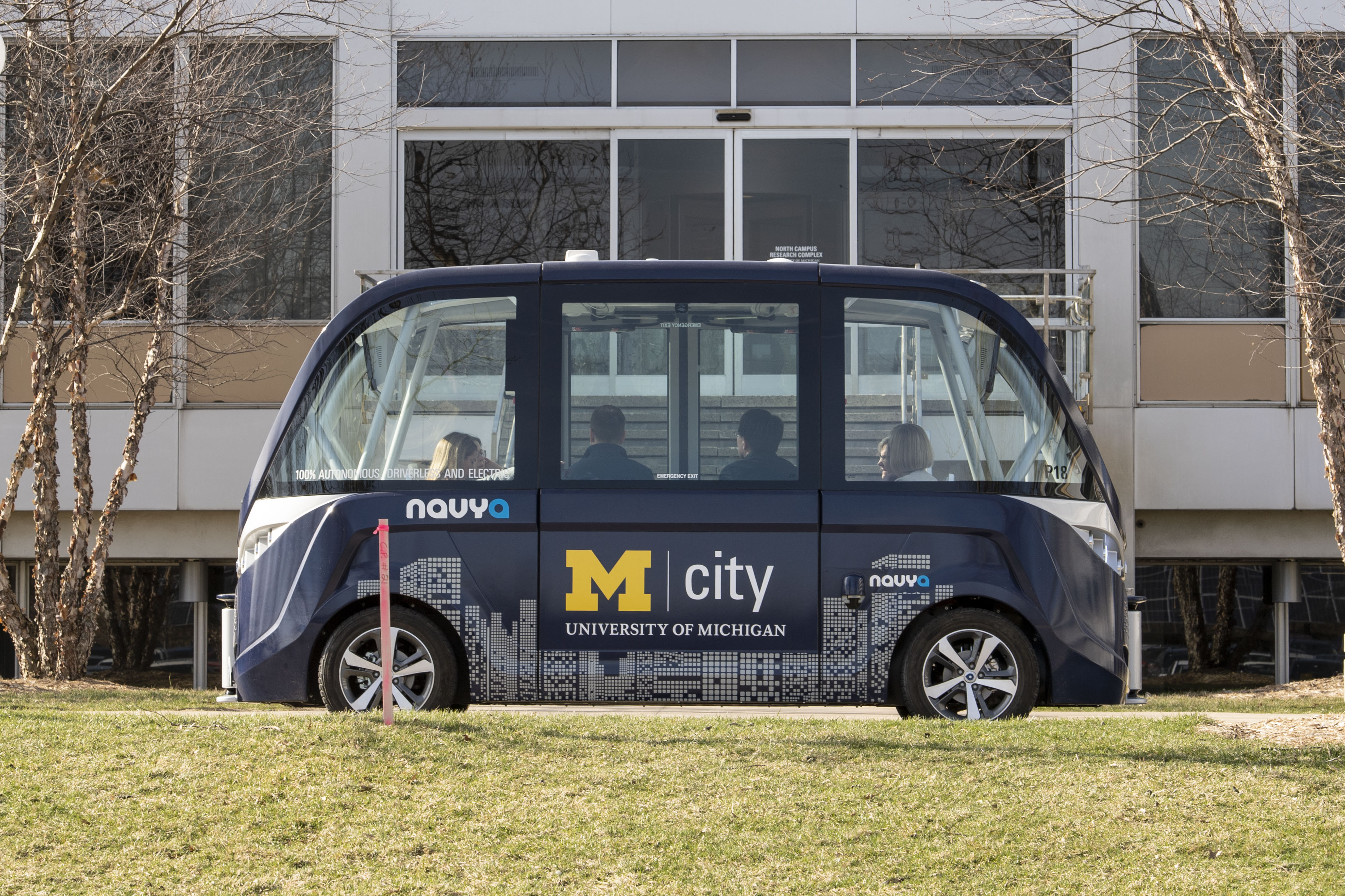 Mcity Driverless Shuttle on its route at the University of Michigan’s North Campus Research Complex. Side view of boxy blue shuttle with large windows and several people visible inside; grass in foreground and building in background
