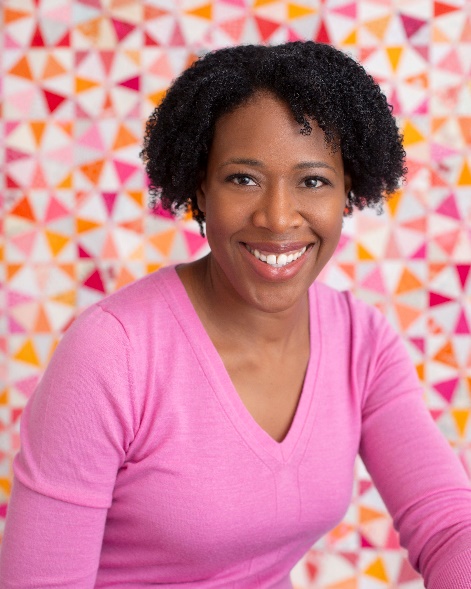 Nicole Neblett, Ann Arbor, Michigan Medium-dark-skinned woman wearing pink top with bobbed curly black hair in front of a colorful backdrop