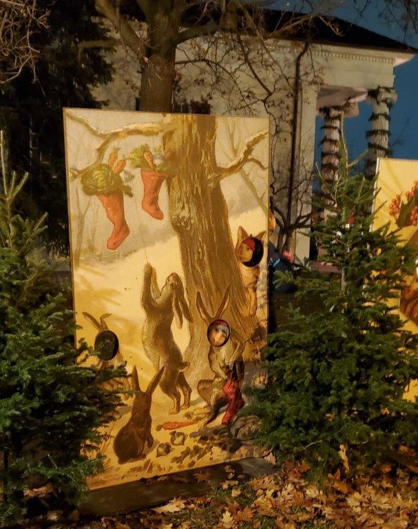 People pose behind a life-size postcard at Holiday Nights in Greenfield Village Two people stick their faces through holes in a life-size holiday postcard among evergreens