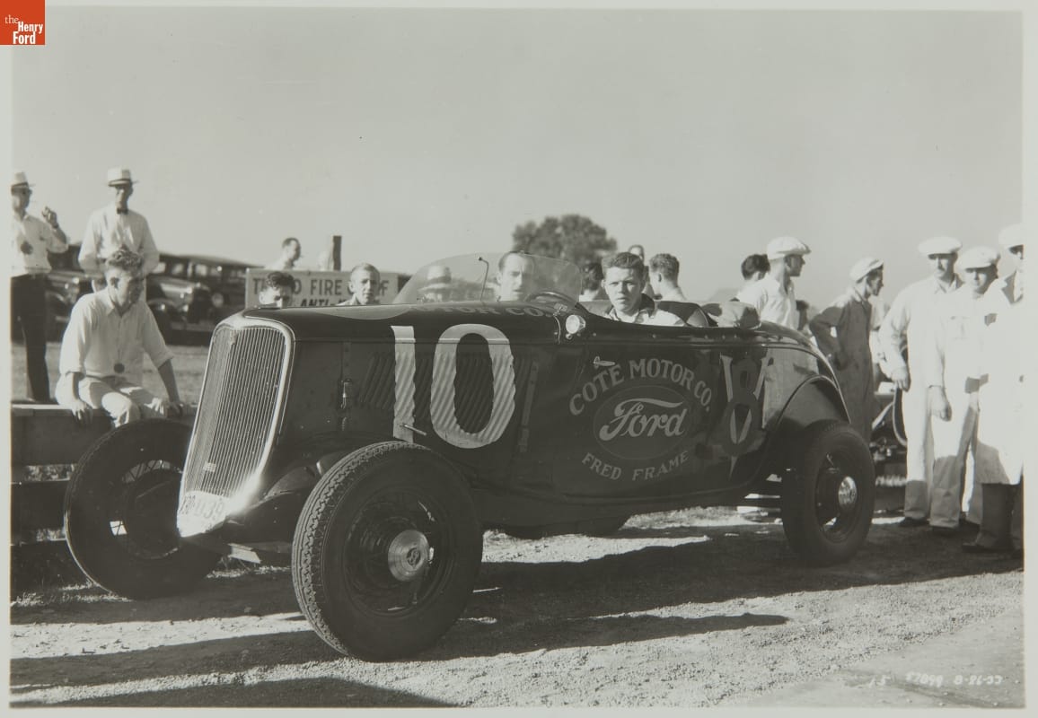 Fred Frame Driving for Cote Motor Company, Elgin National Auto Race Stock Cars, August 1933 Man sits in early, open racecar with other cars and people in the background