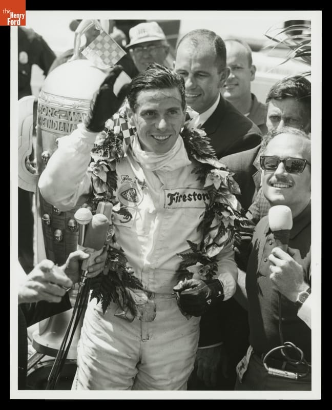 Jim Clark after Winning the 1965 Indianapolis 500 Race Man in jumpsuit and gloves with wreath around neck waves, surrounded by a crowd