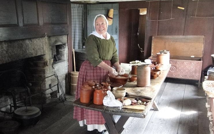 Cindy Melotti preparing food at Daggett Farm in Greenfield Village Woman works at a rustic table in a kitchen with wooden walls and large rustic fireplace