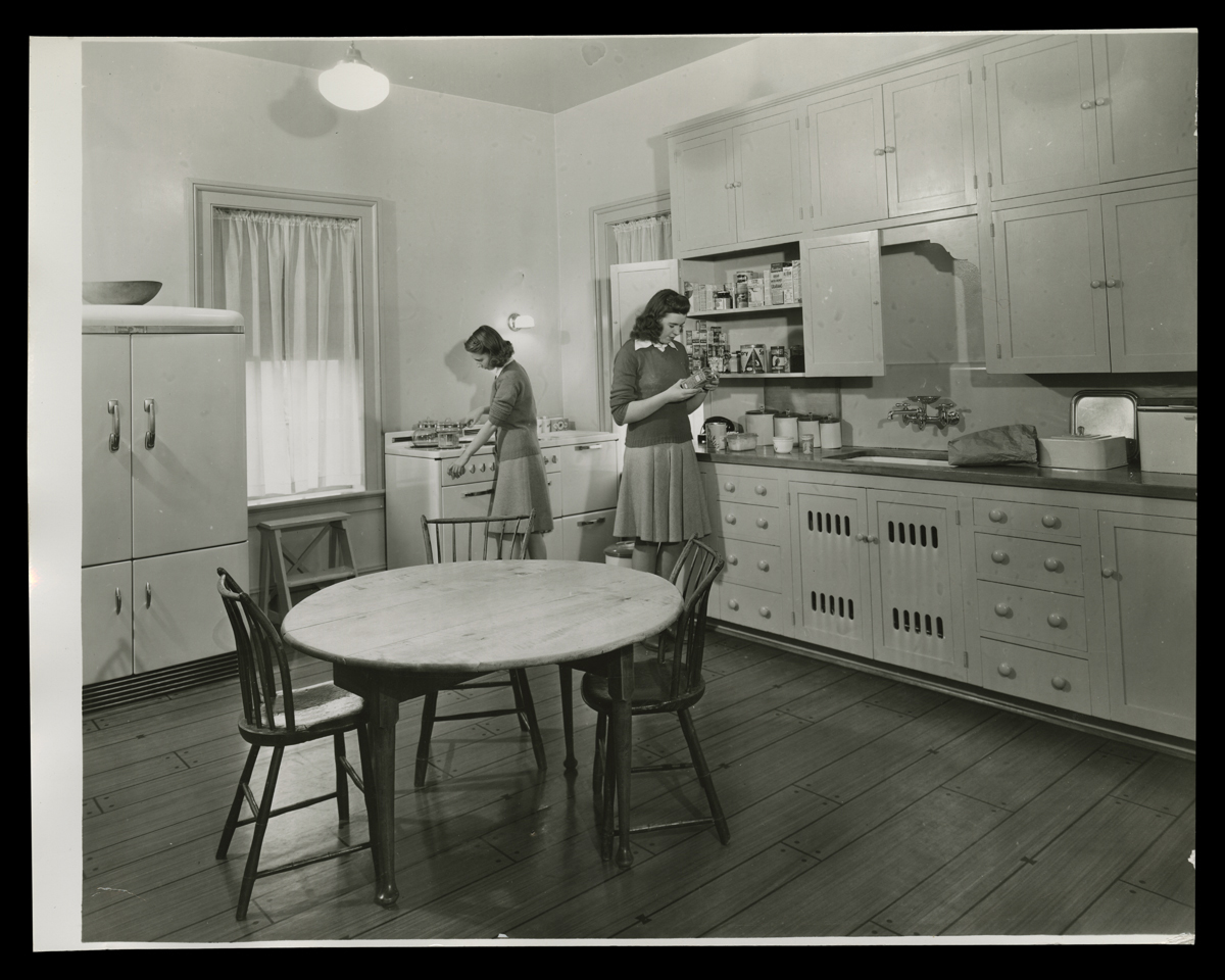 Two young women in a kitchen, one looking at a package from a cabinet and the other at a stove