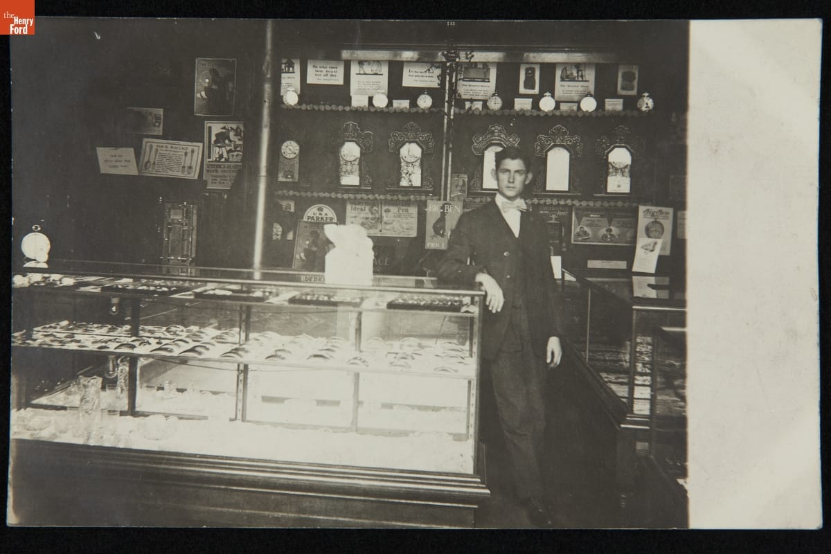 Store Salesman with His Wares, Possibly Urbana, Ohio, 1907-1915 Man in suit and bowtie stands in store with arm on display case and clocks on shelves behind him