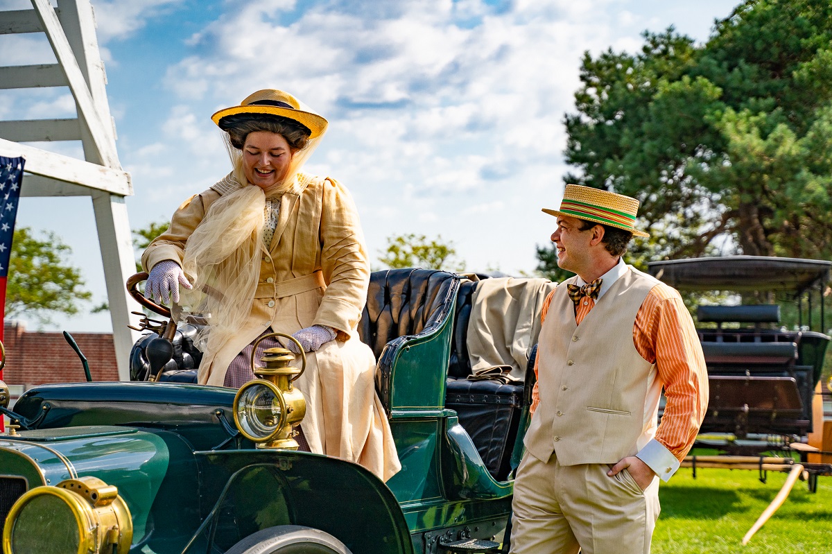 A woman takes the wheel at Old Car Festival 2021 Woman in yellow coat and hat sits behind the wheel of an open car as a man in a vest and straw boater hat stands nearby