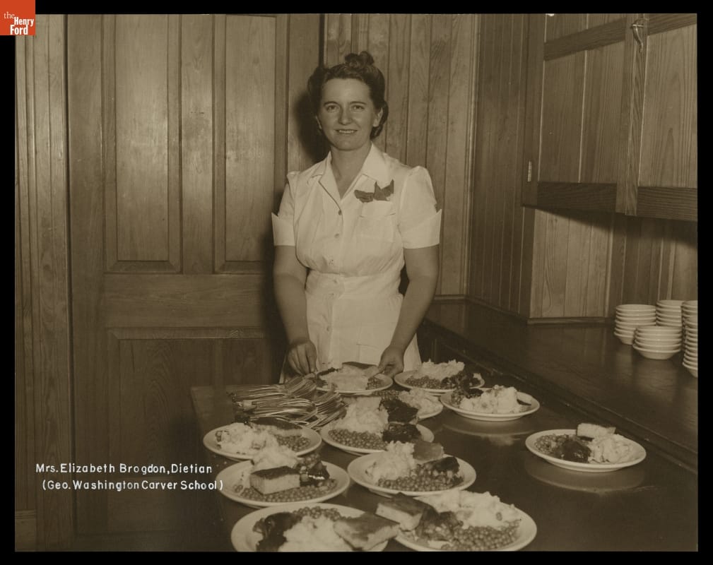 Elizabeth Brogdon, Dietitian at George Washington Carver School, Richmond Hill, Georgia, circa 1947 White woman stands behind table laden with plates of food in wood-paneled room