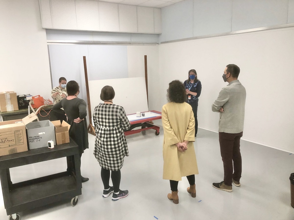 One of our first in-person meetings in the new studio Group of people wearing masks stand in a circle in the corner of a room with several carts filled with boxes nearby